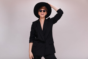 Portrait of stylish lady in sunglasses and wide brimmed hat. Cool young woman in black jacket and pants poses and smiles on isolated background