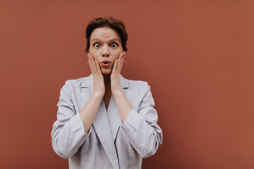 Brown eyed woman in grey suit surprised looking into camera. Short-haired pretty girl in oversize jacket poses on isolated background