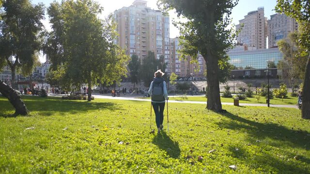 Healthy and active. Mature woman with walking poles walks outdoors on lawn in city park in sunny weather. Cheerful elderly lady in green park working out nordic walking. Scandinavian sticks.