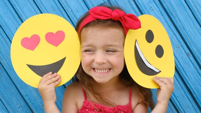 A little girl covers her face with a cardboard smiley face with happy and loving faces . World Emoji Day. An anthropomorphic smiley face.  