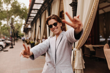 Positive woman in grey jacket and sunglasses showing peace signs outside. Cheerful dark-haired girl in suit widely siles and walks around city
