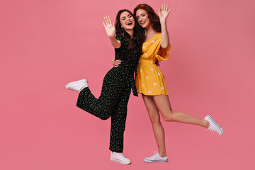 Cheerful women in dresses waving hands in greeting. Two charming girlfriends in beautiful summer clothes rejoice on pink background