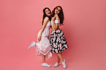 Curly ladies in midi dresses pose on pink background. Charming girls with bright makeup hugging,...
