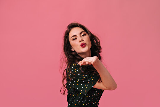 Curly lady with bright lipstick blows kiss on isolated background. Cute girl in dark polka dot dress beautifully and womanly poses in good mood