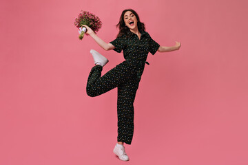 Happy woman holding bouquet and jumping on pink background. Cutie in black polka dot jumpsuit moves...
