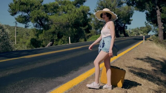Footage of 20s age One woman with a straw hat, jean shorts, knit t-shirt  and yellow suitcase hitchhiking by the country roadside