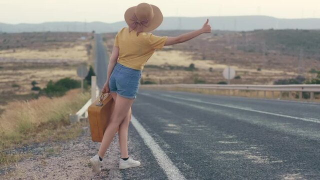 Footage of 20s age One woman with a straw hat, jean shorts, Yellow t-shirt  and yellow suitcase hitchhiking by the country roadside on sunset.