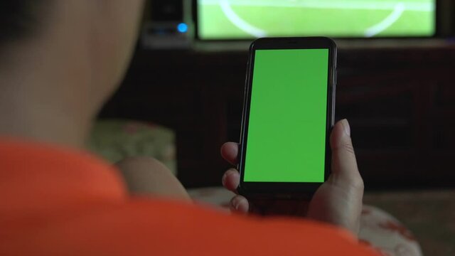 Man relax on couch watching tv and using green mock-up screen smartphone. Close up view.