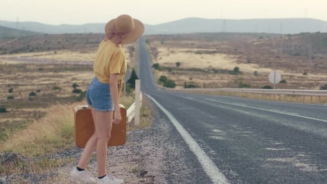 Footage of 20s age One woman with a straw hat, jean shorts, Yellow t-shirt  and yellow suitcase hitchhiking by the country roadside on sunset.