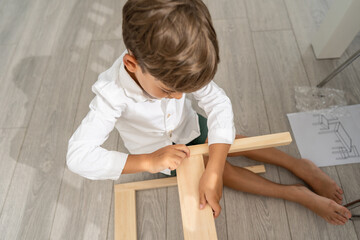 Fototapeta na wymiar Small caucasian boy four years old playing with self assembly chair or table on the floor at home in day - little child assembling furniture alone childhood craft concept top view copy space