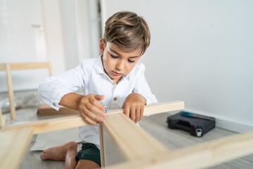 Small caucasian boy four years old playing with self assembly chair or table on the floor at home in day - little child assembling furniture alone childhood craft concept front view copy space