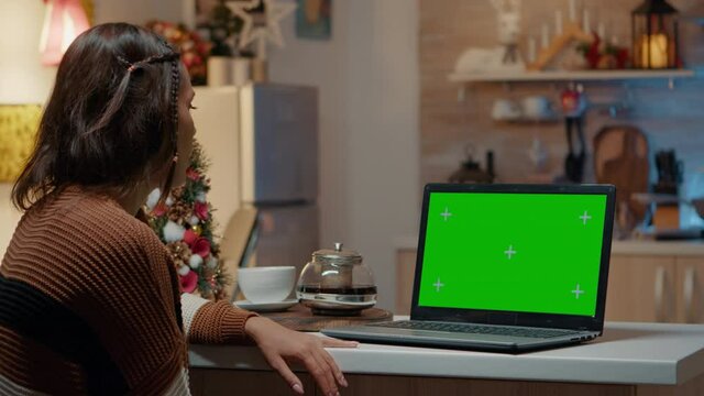 Young adult looking at green screen on laptop in christmas decorated kitchen. Caucasian woman using digital technology device with chroma key mock up electronic concept. Isolated background gadget