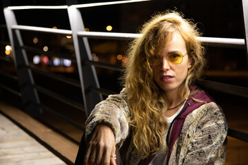 Modern Young blonde woman standing alone in the night time in the city with yellow sunglasses
