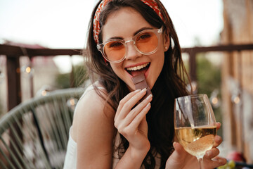 Young girl eats chocolate and holds champagne glass. Charming lady in orange sunglasses with chocolate in her hands is smiling at camera