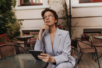 Thoughtful woman in grey suit holds tablet and sits in street restaurant. Charming short-haired...