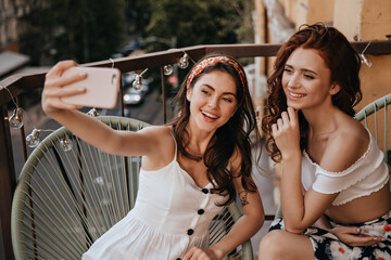 Girls in good humor take selfie on balcony. Curly women with lovely smiles in white modern clothes are photographed on phone