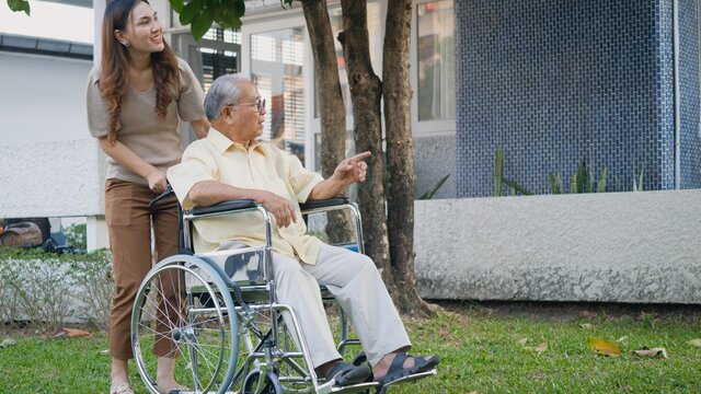 Disabled Senior Man On Wheelchair With Daughter, Happy Asian Generation Family Having Fun Together Outdoors Backyard, Care Helper Young Woman Walking An Elderly Man Smiling And Laughed