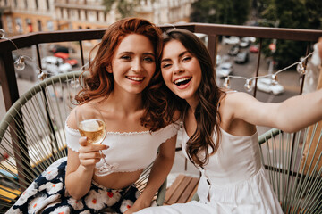 Red haired lady holds champagne glass while brunette woman take selfie. Two cute curly girlfriends...