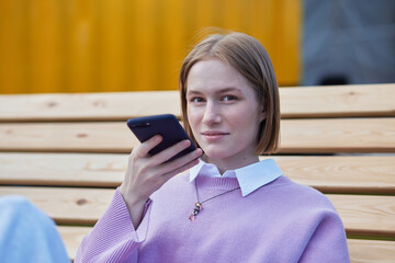 Young smiling woman recording audio message to friend using smartphone sitting on park bench close...