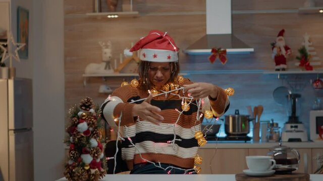 Woman decorating kitchen with festive ornaments while getting tangled in christmas string lights for tree. Young person holding illuminated garland for holiday celebration preparations