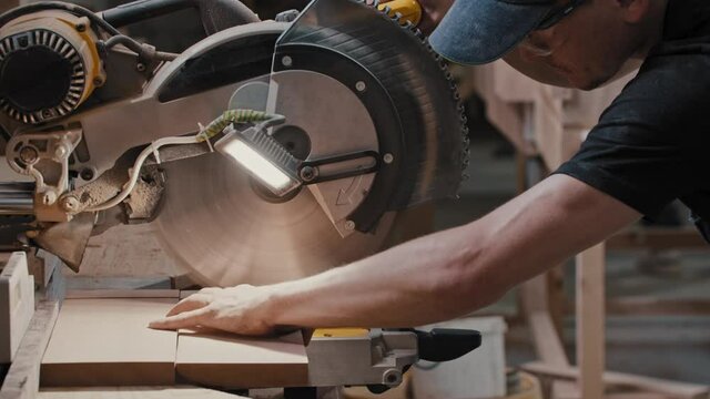 Man in a carpentry workshop cutting a small pieces of the wooden detail using a circular saw