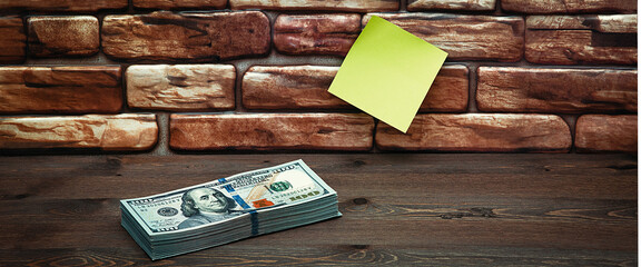 Dollars in one-hundred-dollar bills are stacked on a table near the brick wall. Dollars in a big pile. US dollars. On the brown brick wall there is a place for an inscription, a logo. Color style.