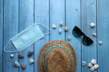 A straw hat, pebbles and shells, sunglasses and a protective mask on a blue wooden background. The concept of recreation and entertainment in the context of the coronavirus pandemic.