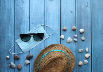 A straw hat, pebbles and shells, sunglasses and a protective mask on a blue wooden background. The concept of recreation and entertainment in the context of the coronavirus pandemic.