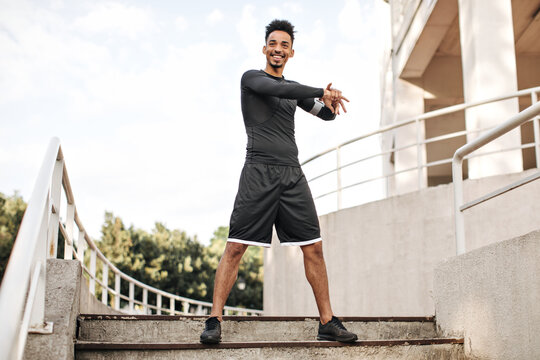 Happy excited brunet dark-skinned man in sport shorts and black t-shirt smiles sincerely, stretches and poses outside at stairs.