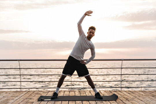 Charming brunette curly dark-skinned man in black shorts and white t-shirt stretches on yoga mat near sea. Young guy works out outside.