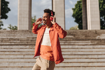 Good-humored man in orange jacket and beige shorts smiles, dancers and listens to music in red headphones outside.