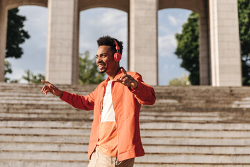 Curly brunet dark-skinned man in orange jacket and stylish t-shirt dances and listens to music in red headphones outside.