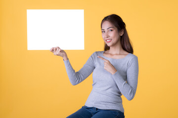 Naklejka na ściany i meble Asian woman in casual clothes holding empty blank board and pointing finger isolated on yellow background