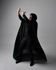 Full length portrait of a  brunette man wearing black shirt, waistcoat and a green velvet cloak.  Standing  action pose isolated  against a grey studio background.