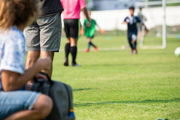 Mother and father watching their son playing football in a school tournament on a clear sky and sunny day. Sport, active lifestyle, happy family and soccer mom, soccer dad concept. © athichoke.pim