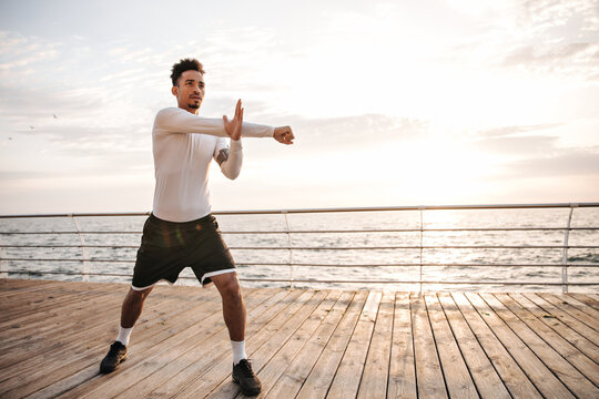 Attractive charming dark-skinned sportsman in black shorts and white long-sleeved t-shirt does ark exercises, stretches and works out near sea.
