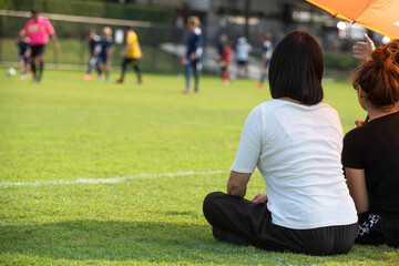 Moms sitting and watching their sons playing football in a school tournament on a sideline with a sunny day. Sport, outdoor active, lifestyle, happy family and soccer mom and soccer dad concepts.