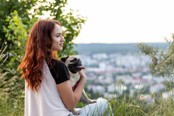 Outdoor portrait of a pug dog and a red-haired woman on a hill at sunset.