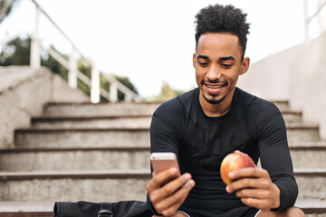 Joyful good-humored brunet dark-skinned sportsman in black long-sleeved t-shirt holds phone, smiles...