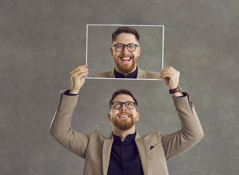 Portrait of a happy smiling bearded young man in glasses with perfect white teeth holding a photo of himself laughing standing isolated on a grey background. Concept of human emotions and dental care