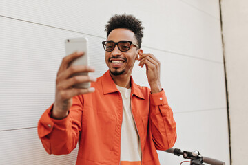 Cheerful curly dark-skinned man in orange jacket and white t-shirt smiles, takes selfie and puts on wireless headphones outside. © Look!