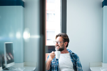 Portrait of happy man looking outside the window and smile drinking a coffee at home ot office - adult caucasian male with beard and glasses in job break activity alone - happy people with eyeglasses