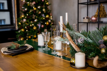 Christmas table setting in rustic style with candles, plates, glasses and eco decors on wooden festive table.New year decorations with fir branches, golden balls, pine cones. Cozy winter holidays.