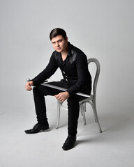 Full length portrait of a  brunette man wearing black shirt and gothic waistcoat holding sword.  Seated  pose isolated  against a grey studio background.