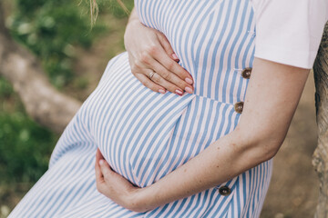 Close-up of the belly of a pregnant girl. A young mother strokes her stomach with her hands. Pregnant woman in a delicate blue striped dress.