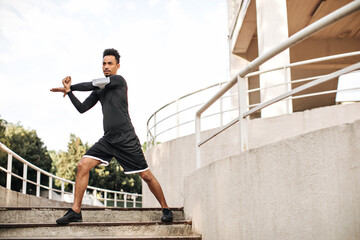 Stylish young man in black sport shorts and long-sleeved t-shirt stretches and works out outdoors on stairs.
