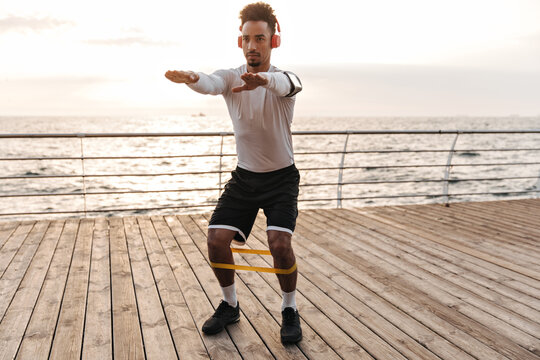 Self-confident young man in black shorts and white t-shirt squats with fitness rubber and listens to music in headphones near sea.