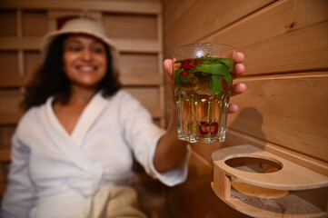 Soft focus on a glass with water with mint, berries and cherries in the hands of a blurred woman relaxing in a wooden sauna