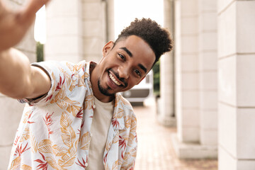 Happy young bearded curly charming man in floral shirt and beige t-shirt smiles sincerely and takes...