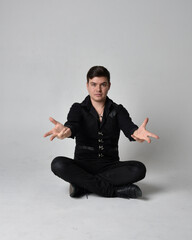 Full length portrait of a  brunette man wearing black shirt and gothic waistcoat.  Seated  pose isolated  against a grey studio background.